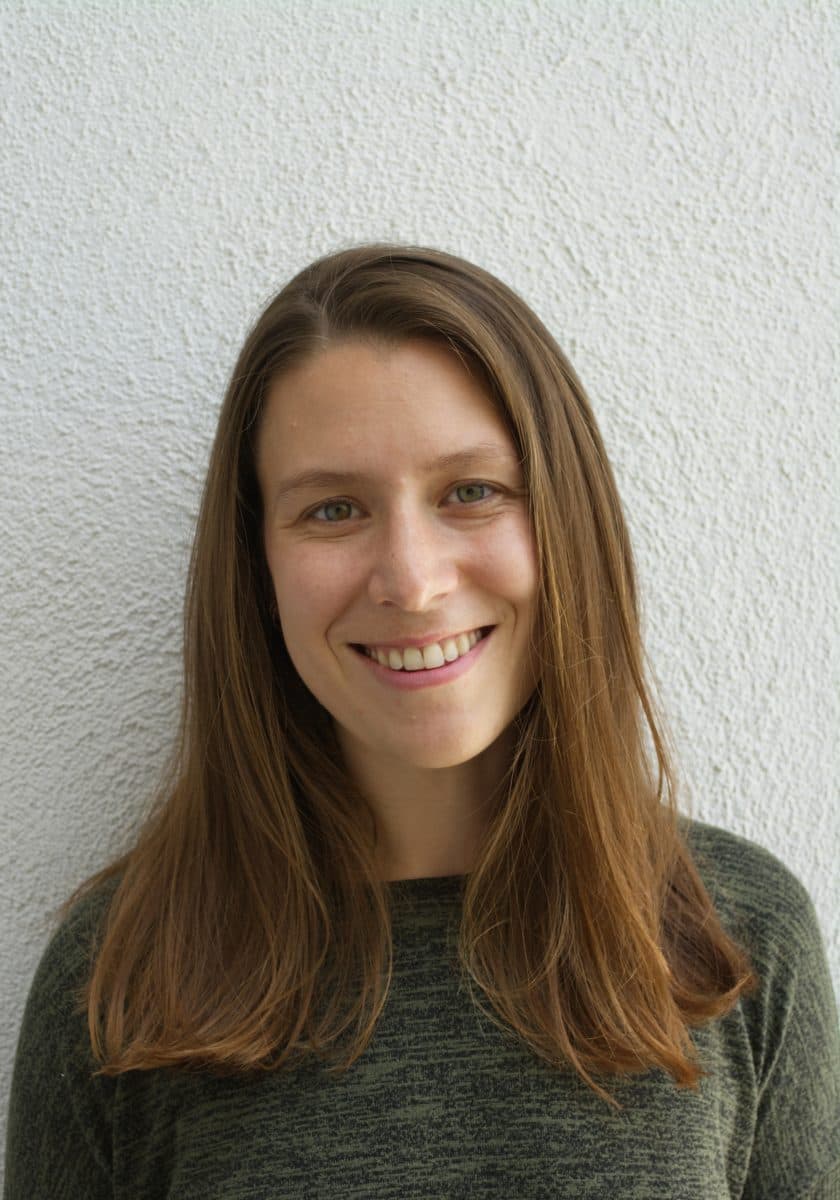 En kvinne med rett, lysebrunt hår og grønn genser smiler foran en hvit, strukturert vegg.