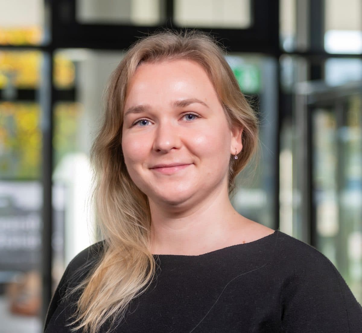 En kvinne med langt, blondt hår og svart topp står innendørs og smiler til kameraet mot en uskarp bakgrunn.