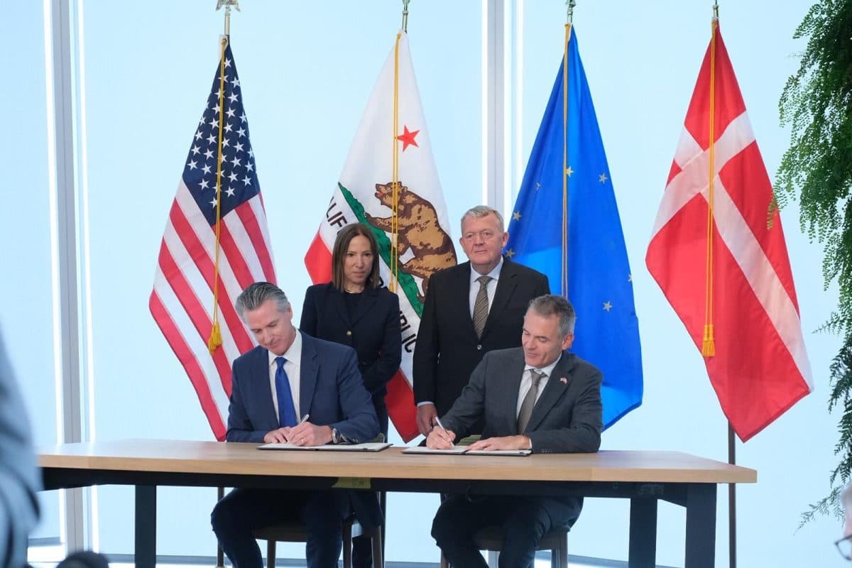 To menn sitter og signerer dokumenter ved et bord, mens to personer står bak dem. I bakgrunnen ser vi flaggene til USA, California, EU og Danmark.