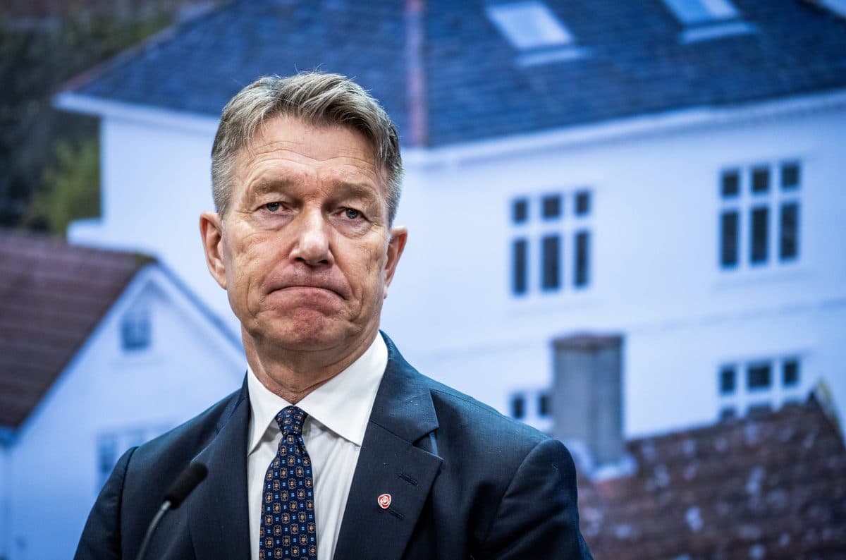 En mann i dress og slips står foran en uskarp bakgrunn med hvite bygninger og ser alvorlig ut.