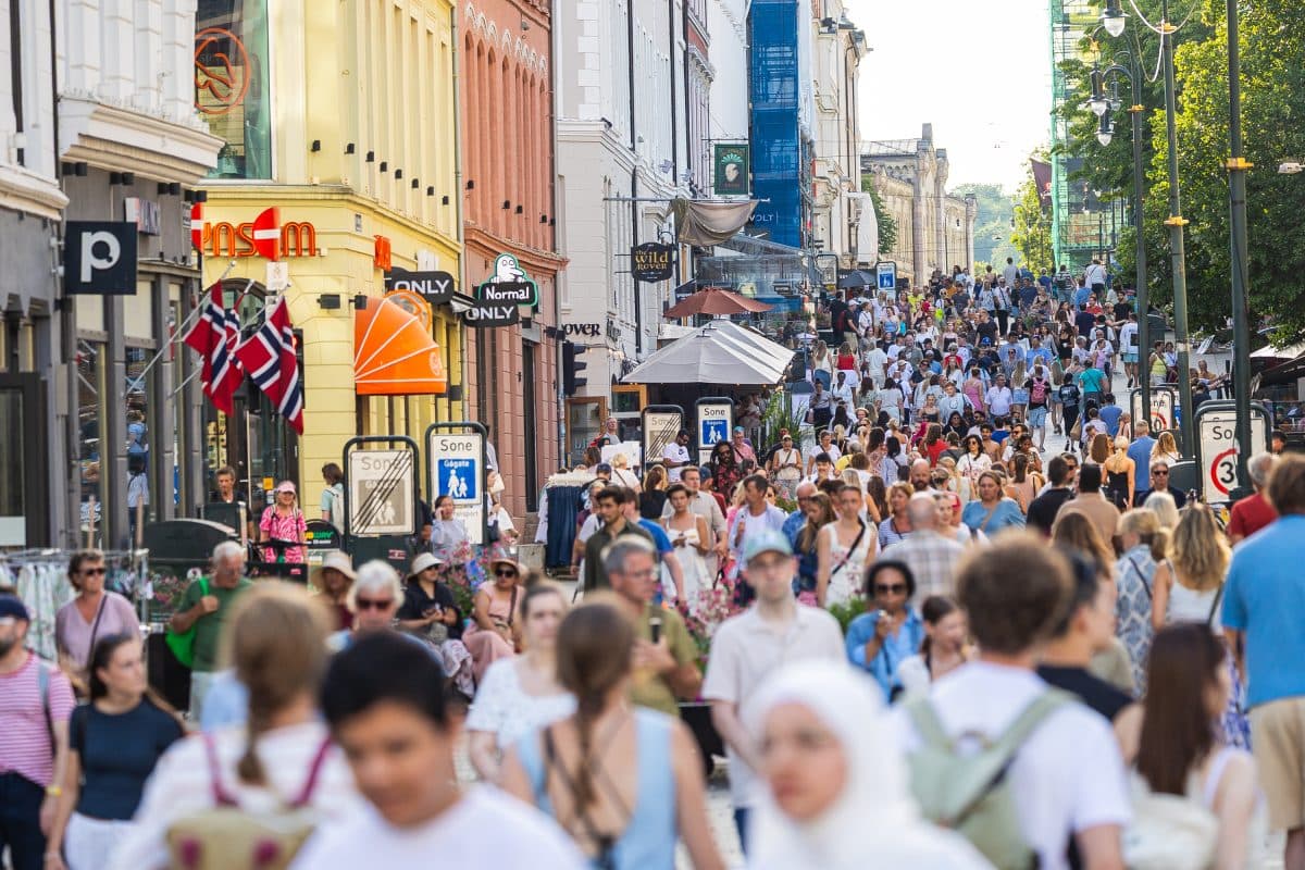 En stor folkemengde går langs en travel bygate med butikker, flagg og skilt på en solfylt dag.
