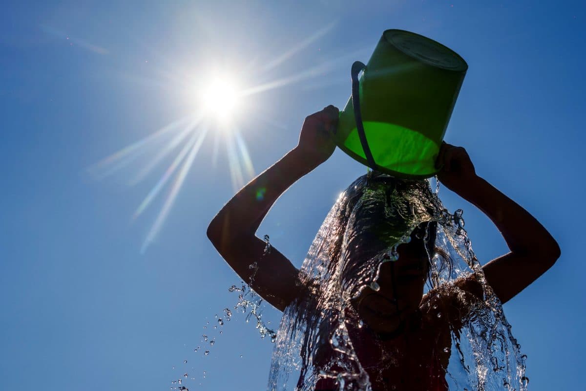 En person heller vann fra en grønn bøtte over hodet under en strålende sol, med vannsprut og klar himmel i bakgrunnen.