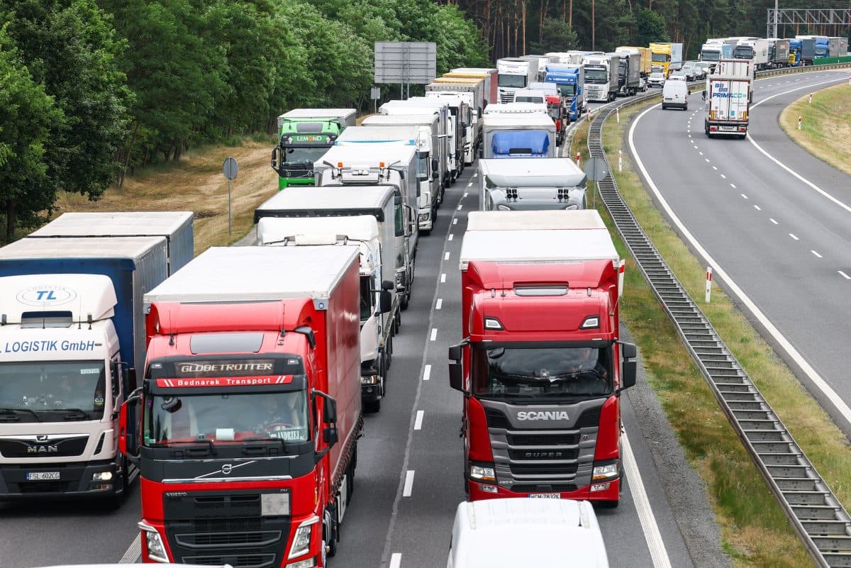En rekke lastebiler står i kø på en flerfelts motorvei, med flere kjøretøy synlige i begge retninger. Trær står langs veien på den ene siden.