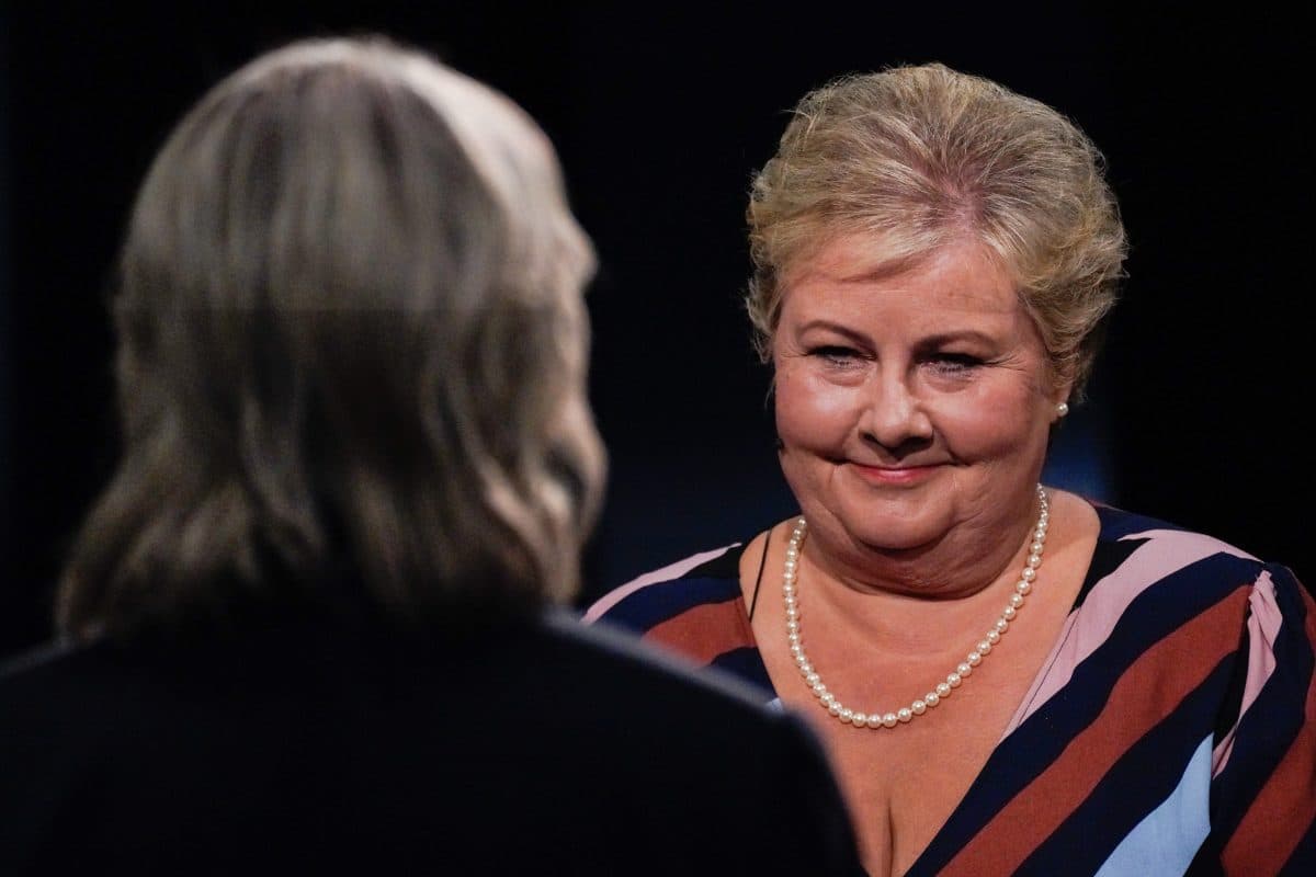 En kvinne med kort, blondt hår, iført stripete kjole og perlekjede, smiler mot en annen person med langt, grått hår, sett bakfra.