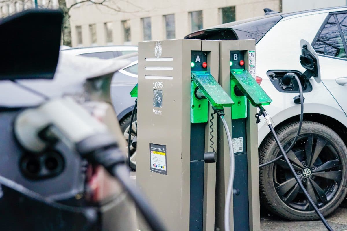 To elbiler er parkert og koblet til en dobbel ladestasjon på en utendørs parkeringsplass.