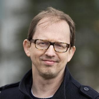 En mann med kort, lysebrunt hår, briller, trimmet bart og skjegg, iført mørk jakke, står utendørs med uskarp bakgrunn.