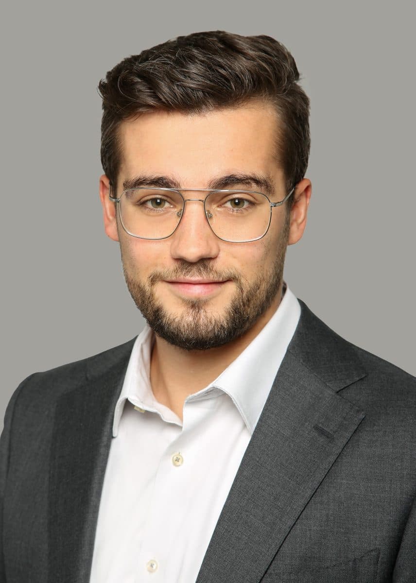 Mann med kort, brunt hår, skjegg og briller, iført hvit skjorte og mørk dressjakke, poserer mot en grå bakgrunn.