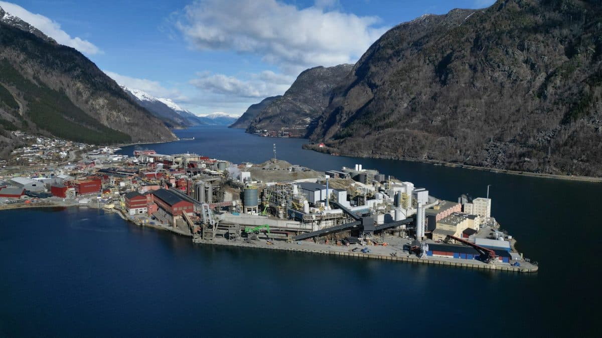 Et industrikompleks med flere bygninger ligger på en halvøy omgitt av vann og fjell under en delvis overskyet himmel.