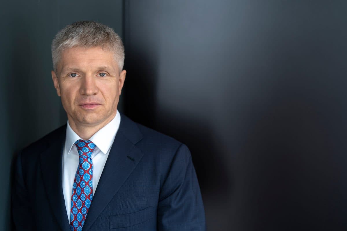 En mann i marineblå dress og mønstret slips står mot en mørk bakgrunn og ser inn i kameraet med et nøytralt uttrykk.