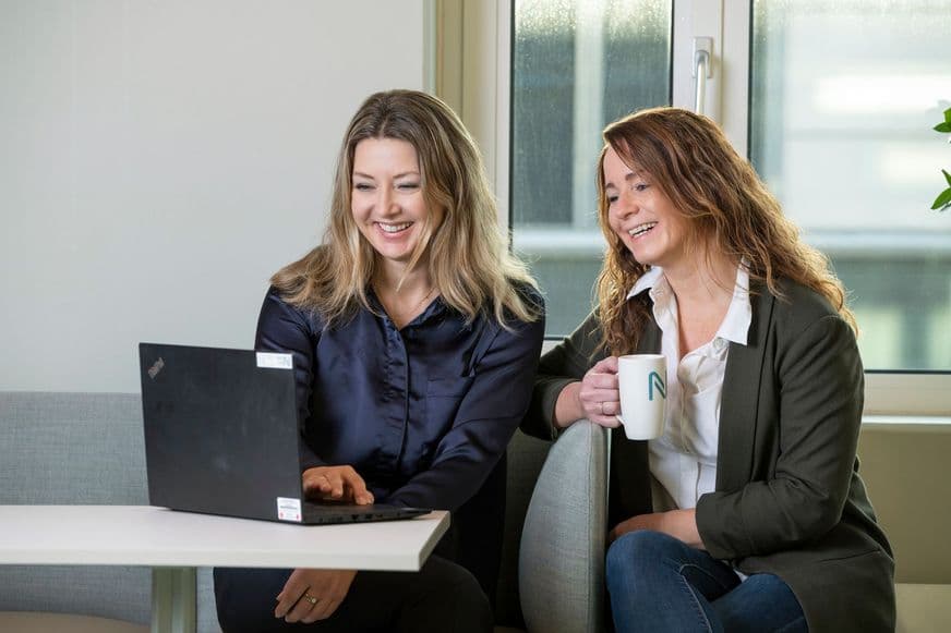 To kvinner sitter i en sofa, den ene med et krus i hånden, begge smiler og ser på skjermen til en åpen laptop som står på bordet foran dem.