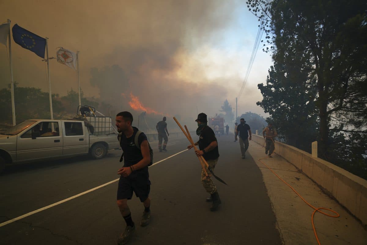 Folk løper og bærer utstyr nær en vei mens tykk røyk og flammer fra en skogbrann sprer seg, med kjøretøy og flagg synlige i bakgrunnen.