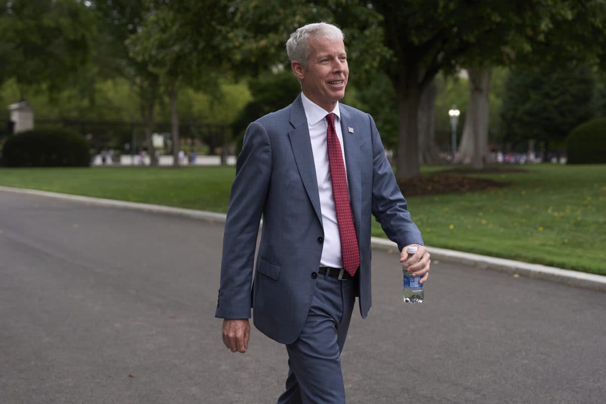 En mann i grå dress med rødt slips går utendørs på en asfaltert sti med en vannflaske i hånden, med grønne trær og gress i bakgrunnen.