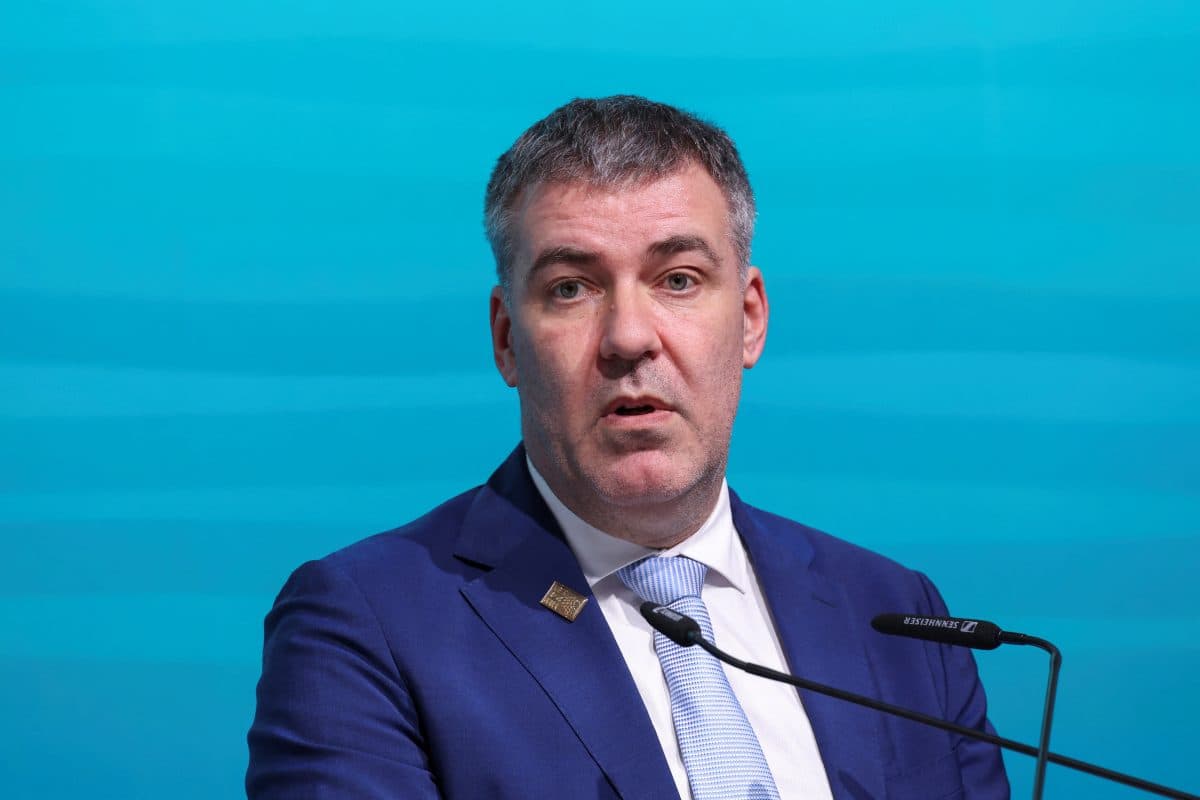 En mann i blå dress og lyseblått slips taler på et podium mot en blå bakgrunn.