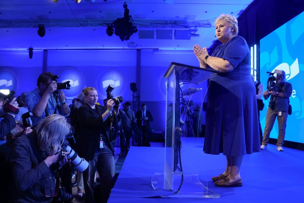 En kvinne i blå kjole taler på et tydelig podium på en scene, mens fotografer tar bilder i et rom med blå belysning.