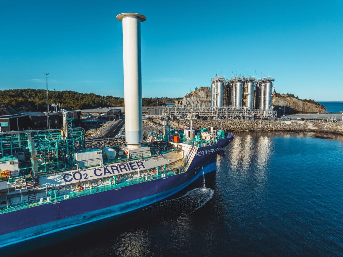 Et skip merket "CO2 CARRIER" ligger til kai ved et industrianlegg med høye lagertanker og skogkledde landområder i bakgrunnen under en klar blå himmel.