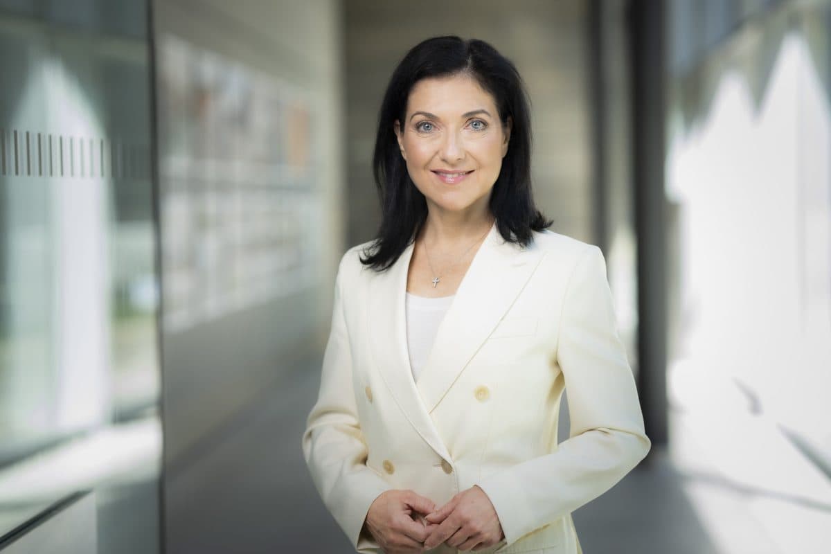 En kvinne med langt, mørkt hår og en kremfarget blazer står i en moderne gang og smiler til kameraet.