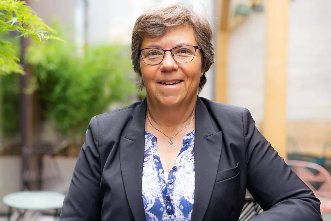 En kvinne med kort, brunt hår og briller, iført en marineblå blazer over en bluse med trykk, smiler mens hun sitter utendørs i en gårdsplass.