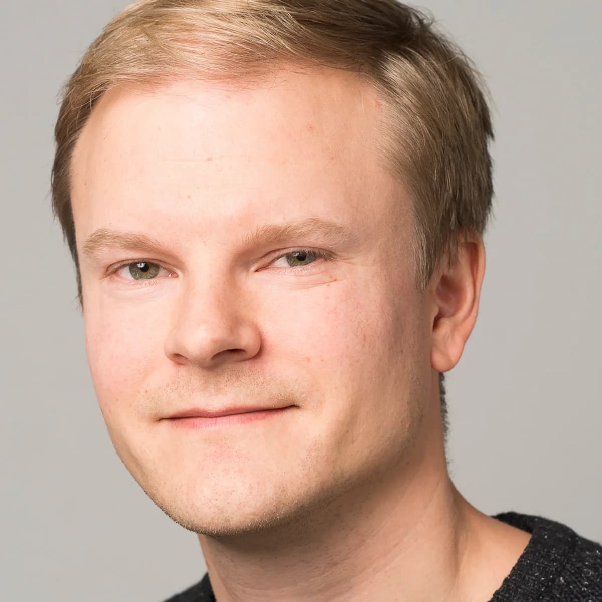 En mann med lys hud og kort, blondt hår ser nøytralt inn i kameraet, iført mørk genser mot en ensfarget, lys bakgrunn.