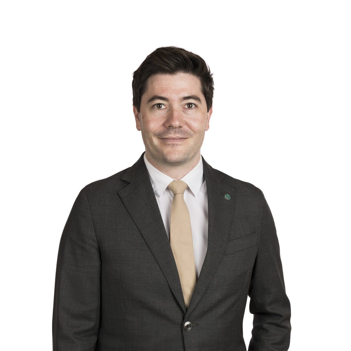 En mann i mørk dress, hvit skjorte og beige slips står vendt fremover mot en enkel, hvit bakgrunn.