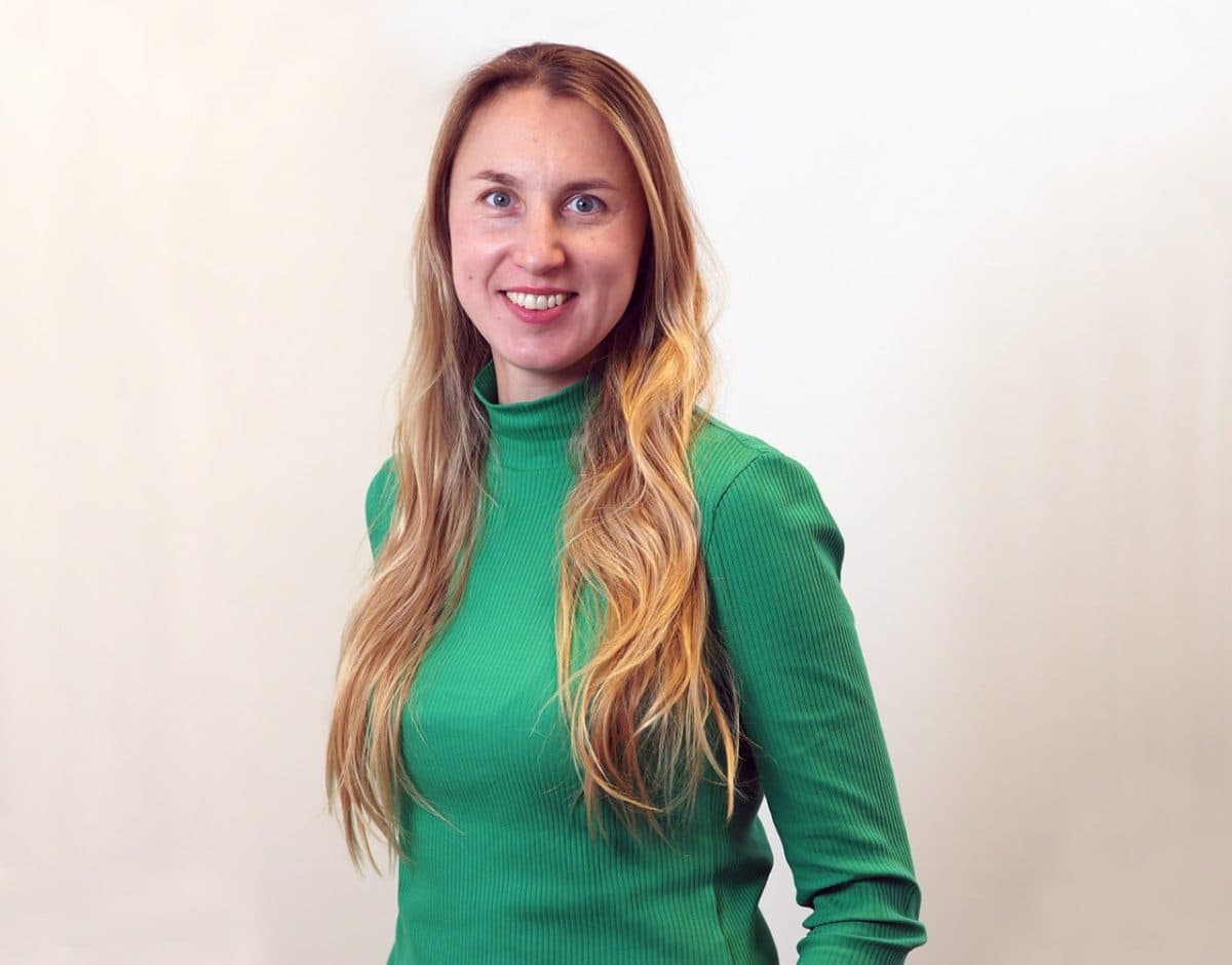 En kvinne med langt, blondt hår og grønn rullekrage står mot en lys bakgrunn og smiler til kameraet.