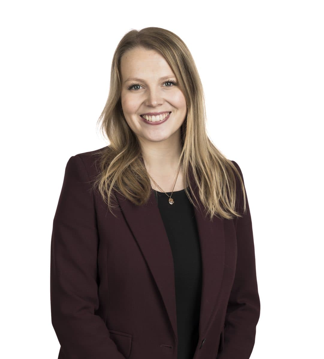 En kvinne med langt blondt hår, iført rødbrun blazer og svart topp, står smilende mot en hvit bakgrunn.