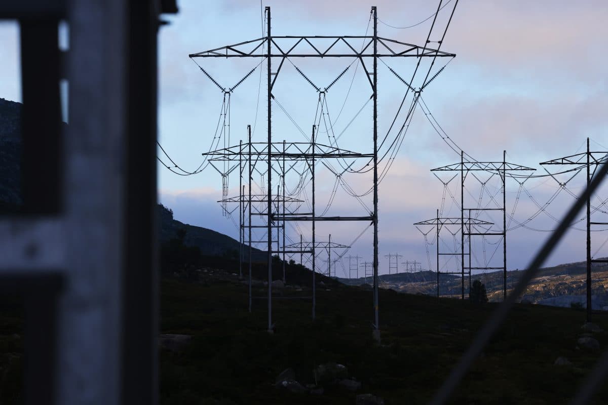 Flere høyspentledninger og master strekker seg over et landlig landskap med åser i bakgrunnen under en overskyet himmel.