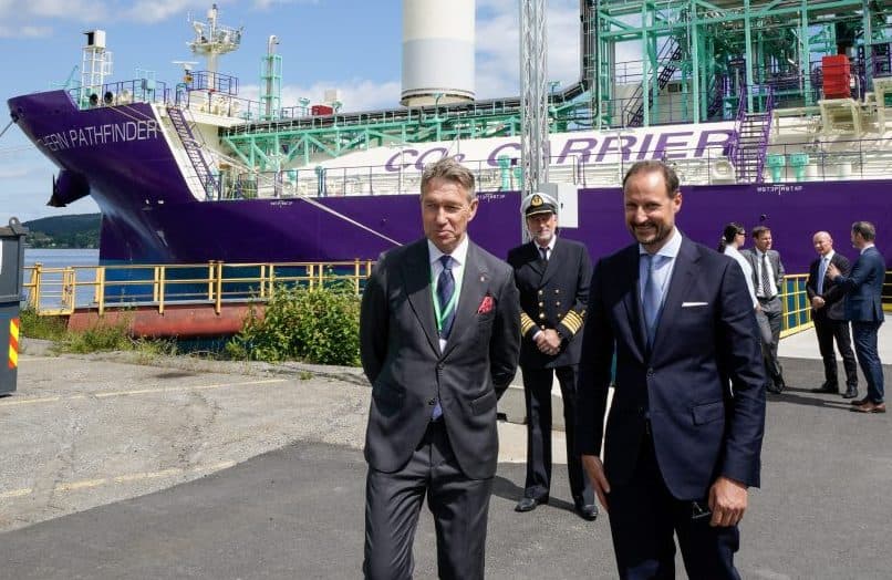 To menn i dress står foran et lilla skip merket "CARRIER", med en uniformert offiser og en annen mann bak seg, på et industriområde.