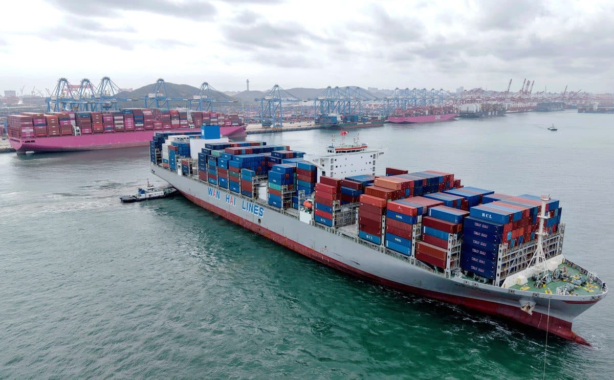 Et stort containerskip lastet med stablede containere seiler i nærheten av en travel havn, med kraner og andre containerskip synlige i bakgrunnen under en overskyet himmel.