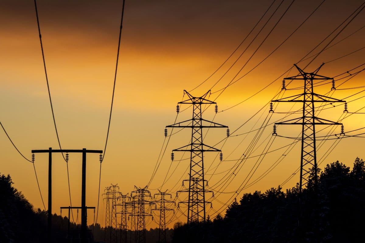 Høyspentledninger og kraftmaster strekker seg over et landskap i solnedgang, i silhuett mot en oransje og gul himmel.