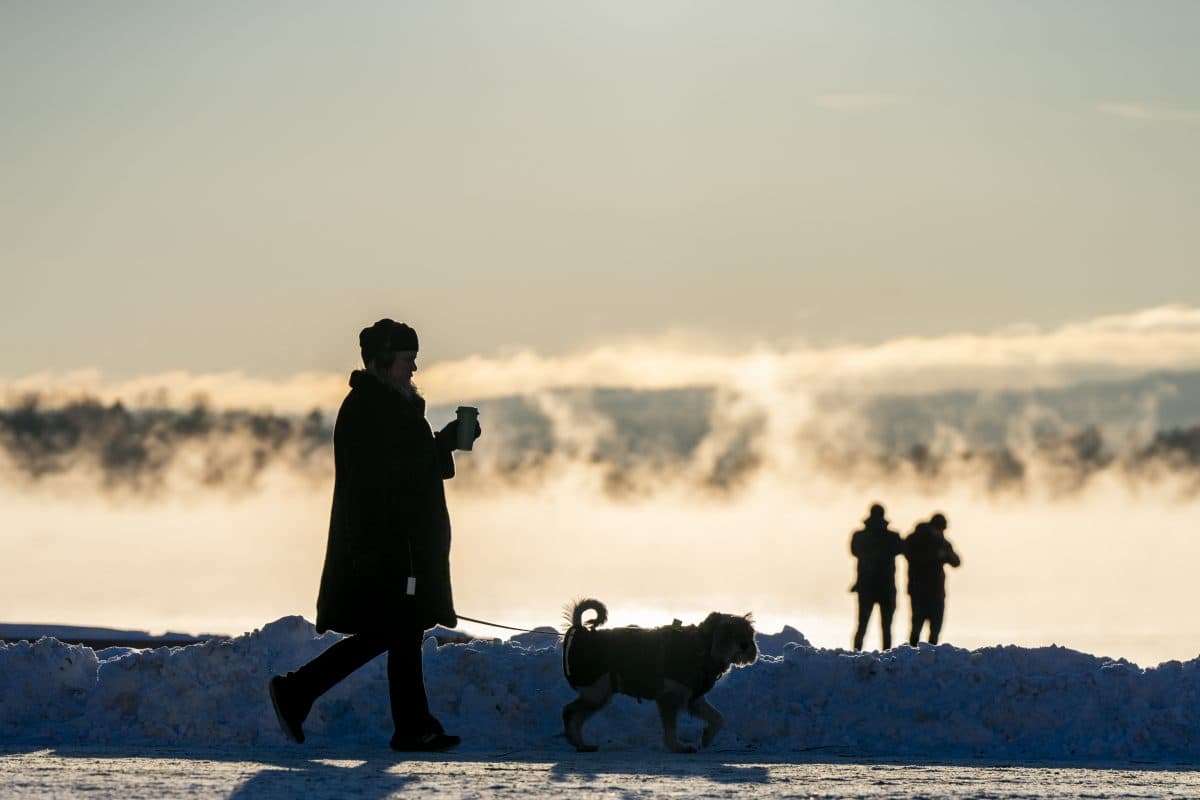 En person går tur med en hund på en snødekt sti mens han holder en drink, med damp som stiger opp fra et vann og to mennesker som står i bakgrunnen.