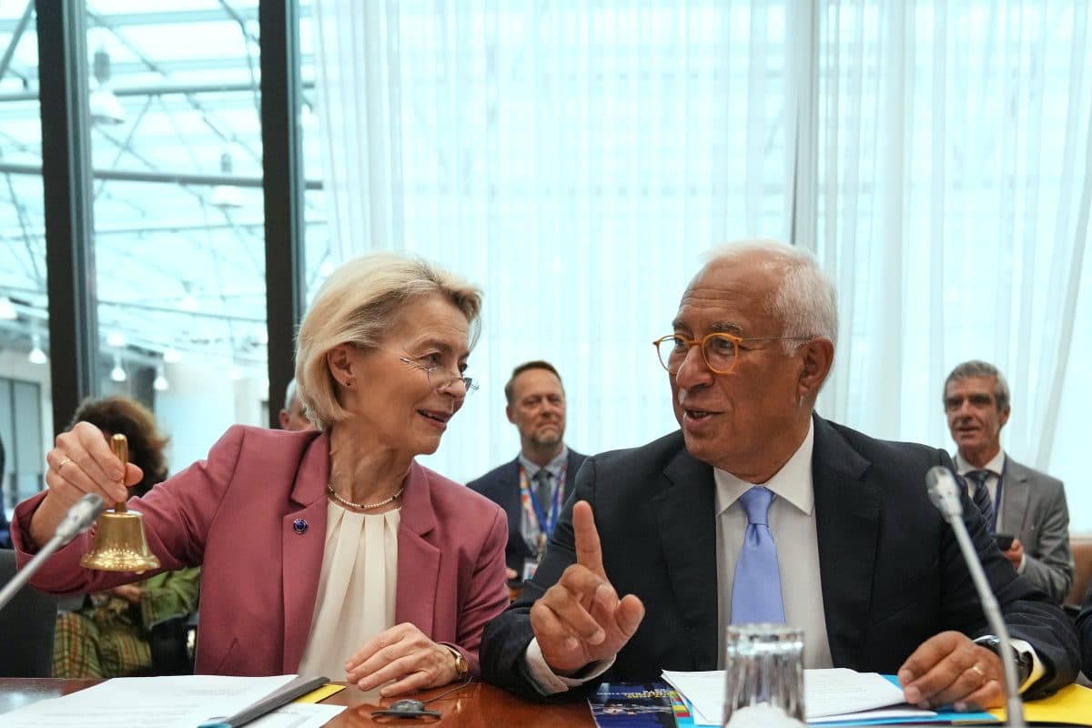 En kvinne med en håndklokke sitter ved siden av en mann i dress som snakker, ved et konferansebord med dokumenter, vann og mikrofoner. I bakgrunnen sitter andre mennesker.