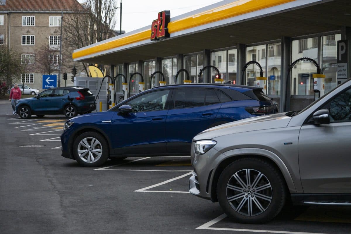 Flere elbiler står parkert og lader ved en ladestasjon utenfor en bygning med gul skilting.