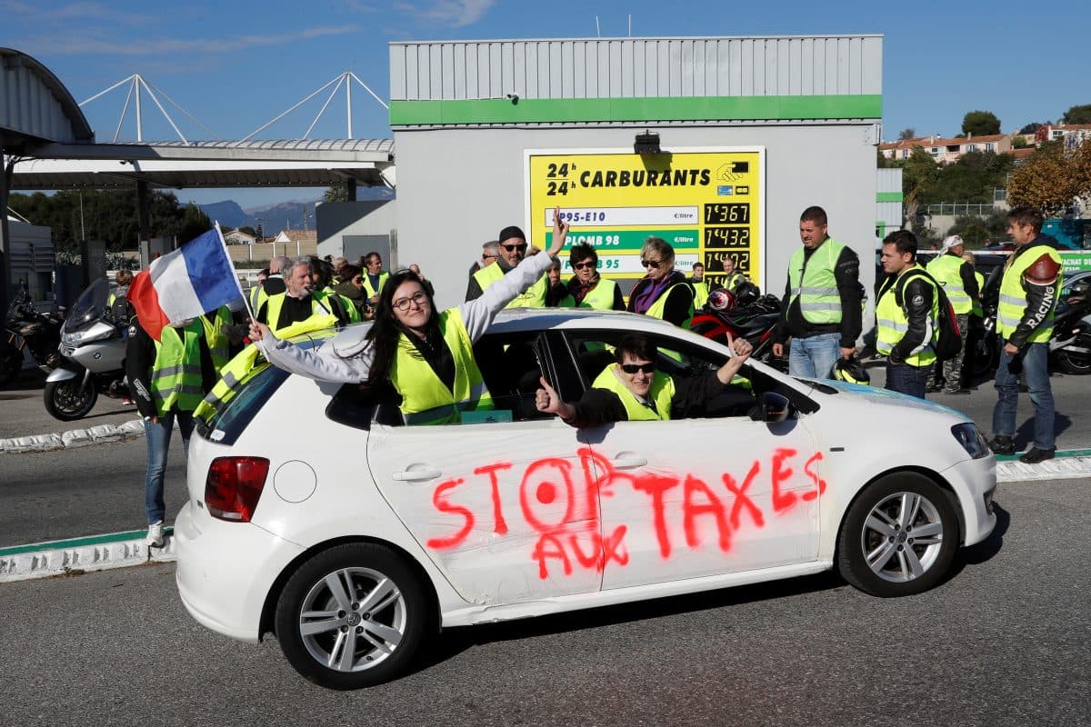 Demonstranter i gule vester samles ved en bensinstasjon; i forgrunnen står en bil med "STOP AUX TAXES" sprayet på siden, og i bakgrunnen holder folk et fransk flagg.
