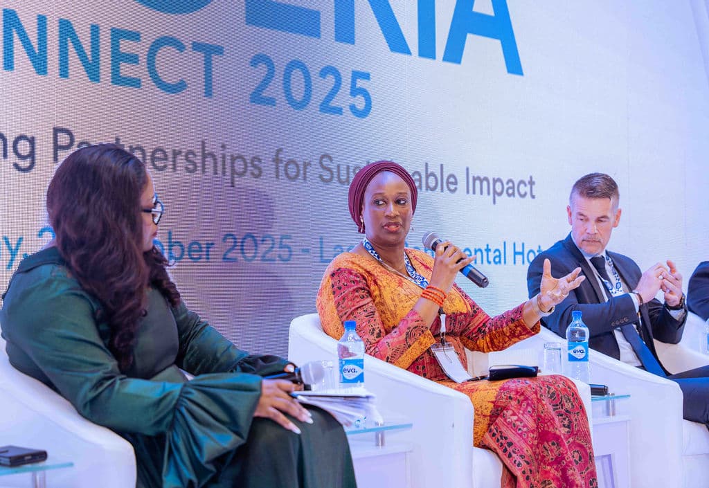 Tre paneldeltakere sitter på scenen på en konferanse, der en kvinne i rød kjole snakker i en mikrofon mens de andre lytter.