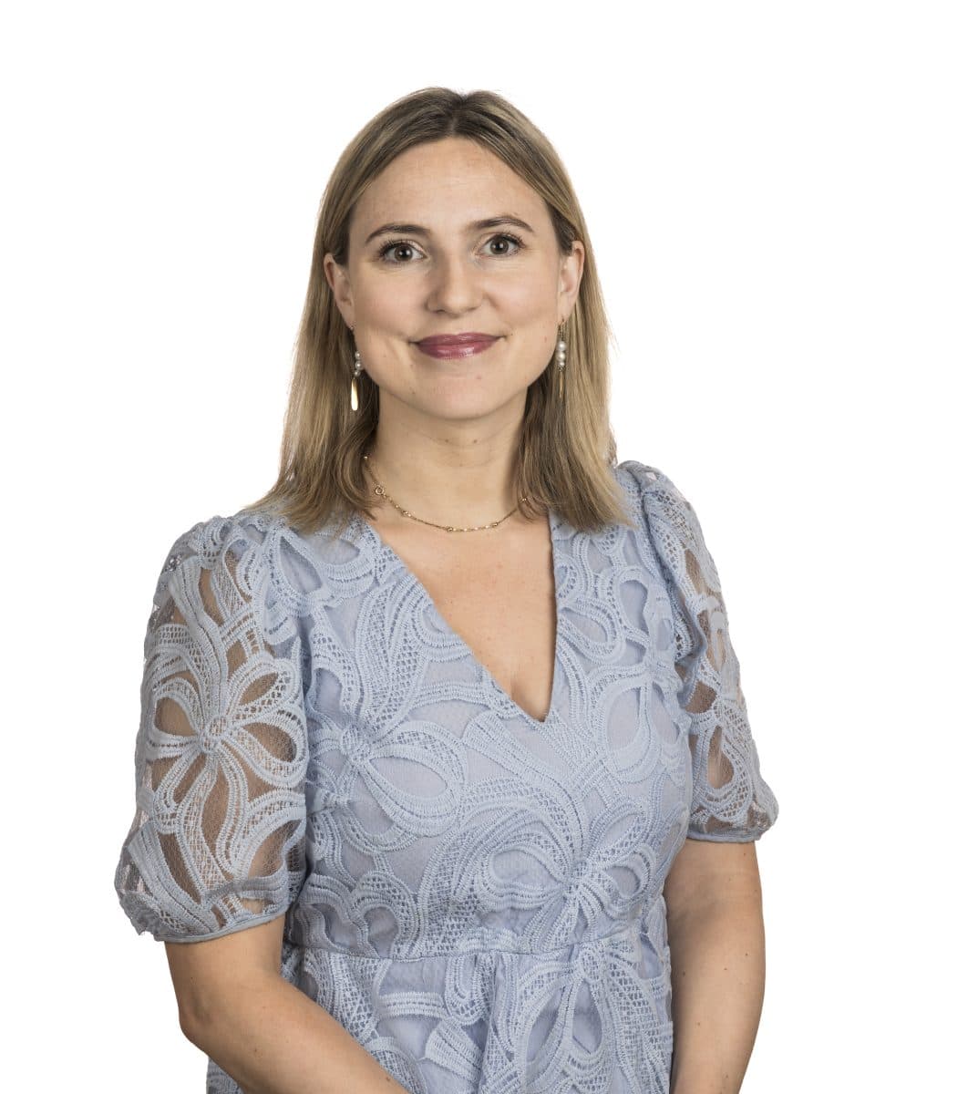 En kvinne med skulderlangt, blondt hår og en lyseblå blondekjole står vendt mot kameraet og smiler lett mot en hvit bakgrunn.