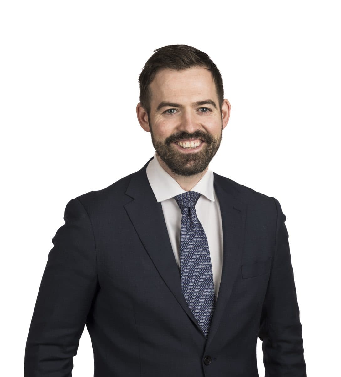 En mann med mørkt hår og skjegg, iført marineblå dress, hvit skjorte og mønstret slips, står smilende mot en vanlig hvit bakgrunn.