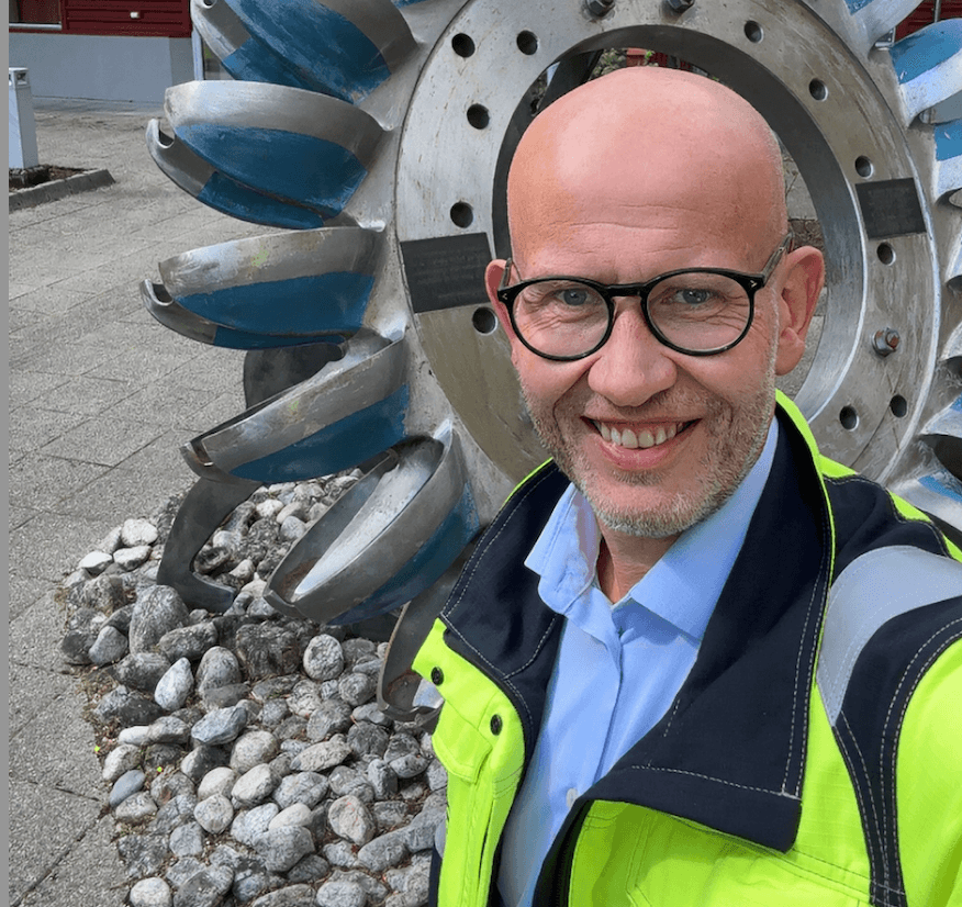 Blid mann med briller og lyseblå skjorte og signalgul arbeidsjakke foran vannturbin ved et kontorbygg