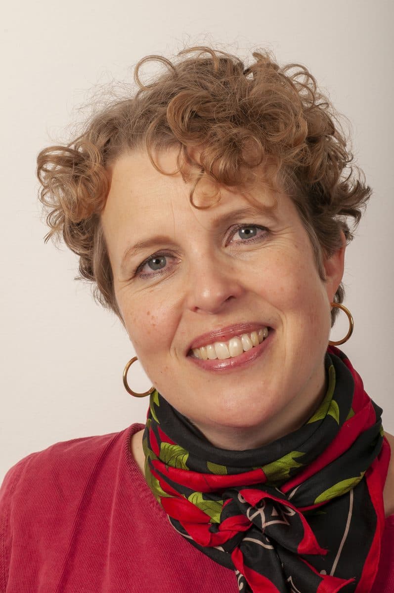 Kvinne med kort, krøllete hår iført øreringer, rød topp og et rødt og svart skjerf, smiler til kameraet foran en ensfarget bakgrunn.