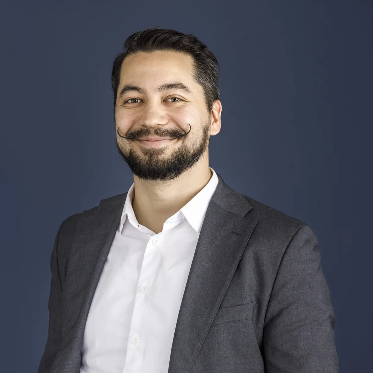 En mann med stylet bart, skjegg og mørkt hår er iført hvit skjorte og grå dressjakke, og smiler foran en ensfarget mørkeblå bakgrunn.