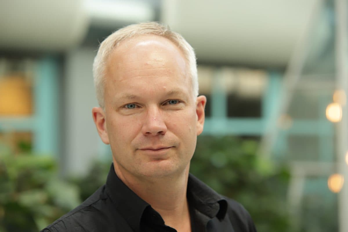 En middelaldrende mann med kort, blondt hår og svart skjorte står innendørs, med en uskarp bakgrunn med vinduer og grønne planter.