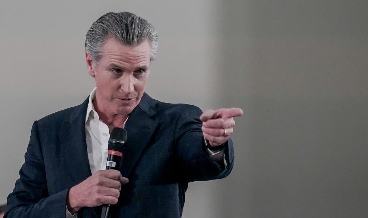 En mann i dress holder en mikrofon i den ene hånden og peker fremover med den andre hånden mot en ensfarget bakgrunn.