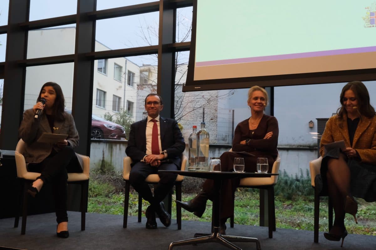 Fire personer sitter på en scene foran store vinduer; en kvinne til venstre snakker i en mikrofon mens de andre lytter, med vannflasker og glass på bordet.