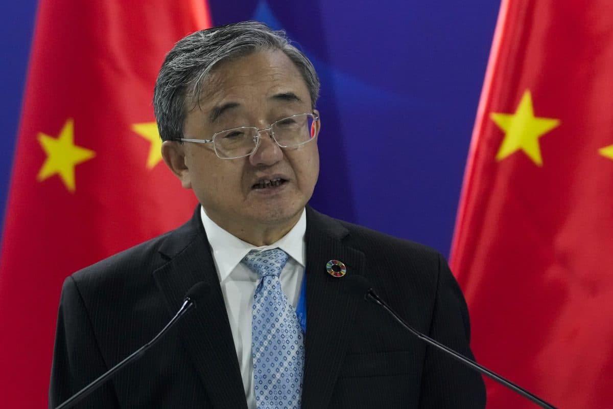 En eldre mann i dress og slips taler på et podium med mikrofoner, med to kinesiske flagg i bakgrunnen.