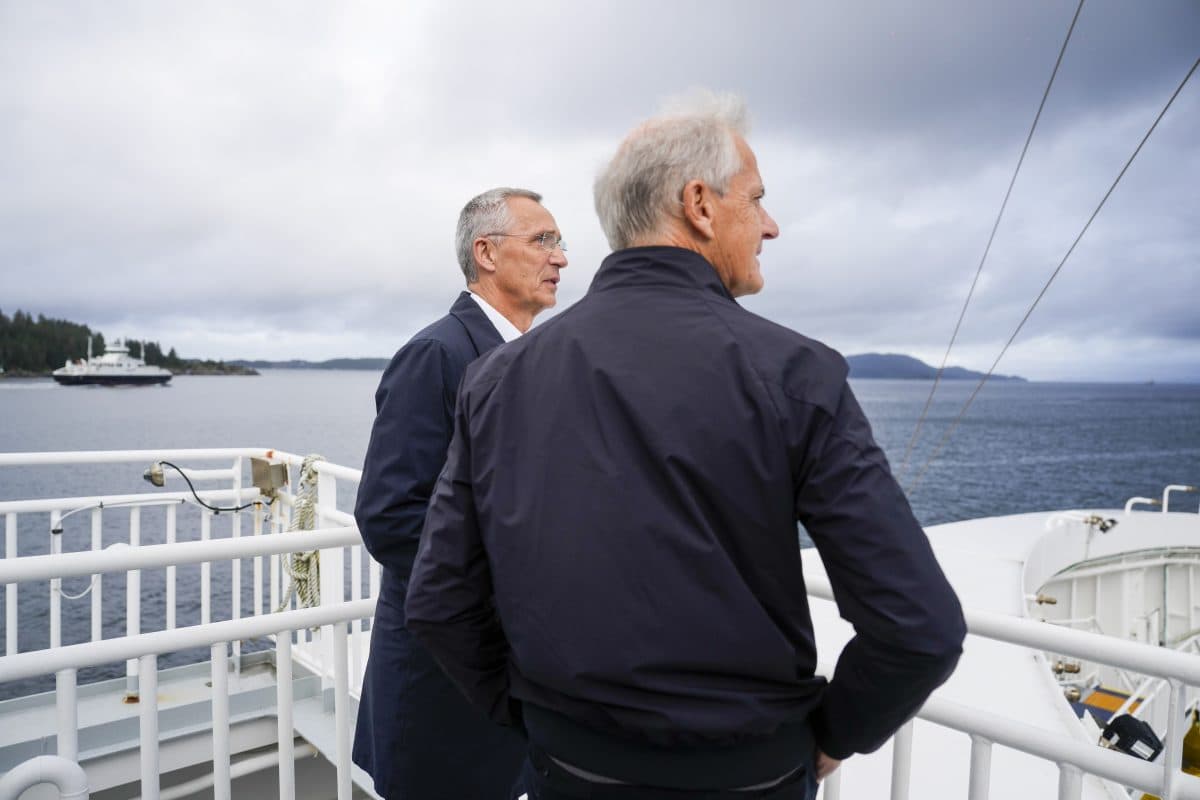 To menn står på dekket av en båt og ser ut over vannet, med land og en annen båt synlig i det fjerne under en overskyet himmel.