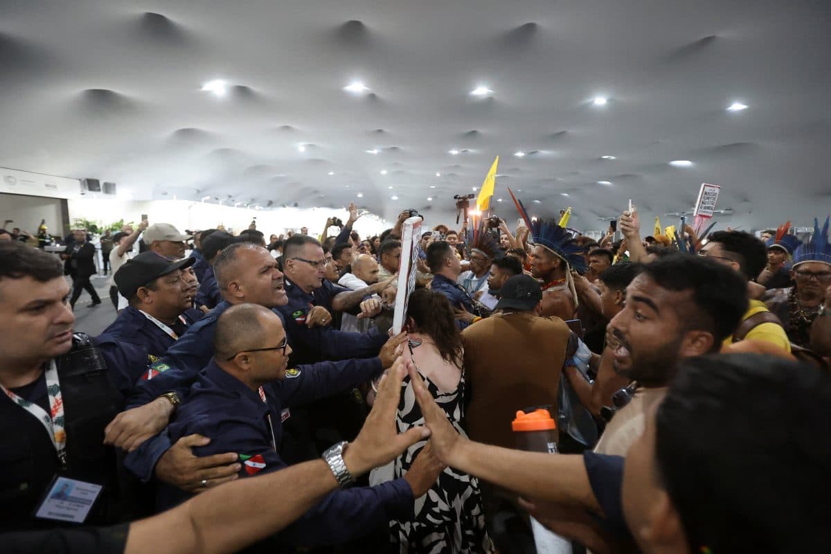 Sikkerhetsfolk og demonstranter støter sammen i et overfylt innendørsrom, der folk dytter og rekker opp hendene, noe som skaper en anspent atmosfære.