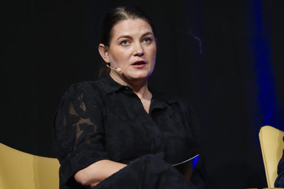 En kvinne med mørkt hår, svart mønstret kjole og hodetelefonmikrofon sitter på en gul stol mot en mørk bakgrunn.