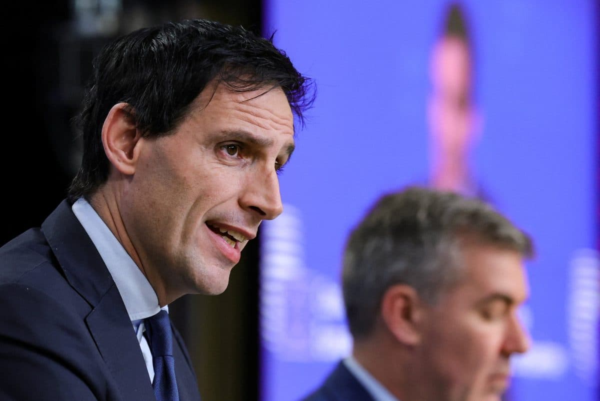 To menn i dress snakker på en pressekonferanse; mannen i forgrunnen snakker, mens mannen i bakgrunnen ser ned.