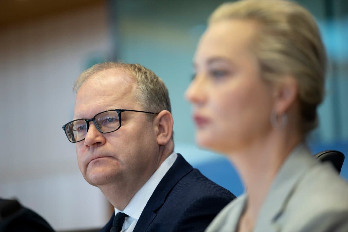 En mann med briller og dress sitter ved siden av en kvinne med blondt hår i en uskarp forgrunn, og begge virker fokuserte i en profesjonell setting.