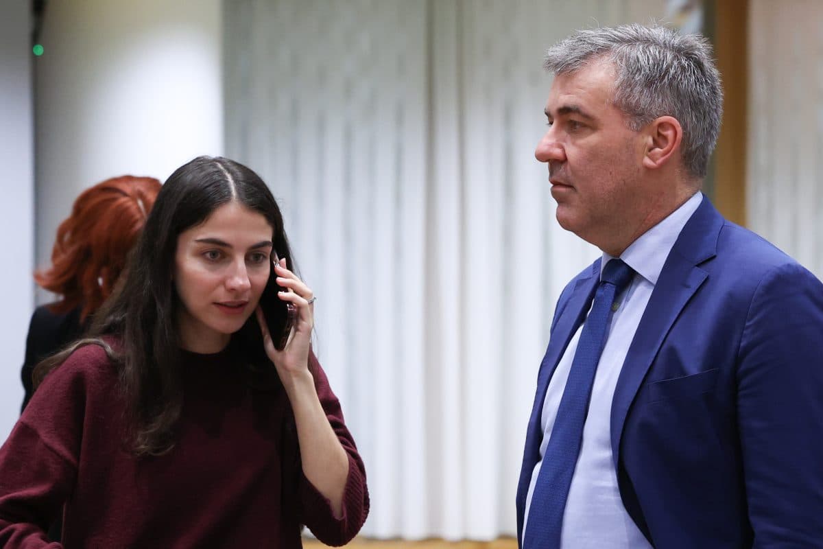 En kvinne snakker i telefon mens hun står ved siden av en mann i blå dress og slips; begge ser ut til å være innendørs på et arrangement eller møte.