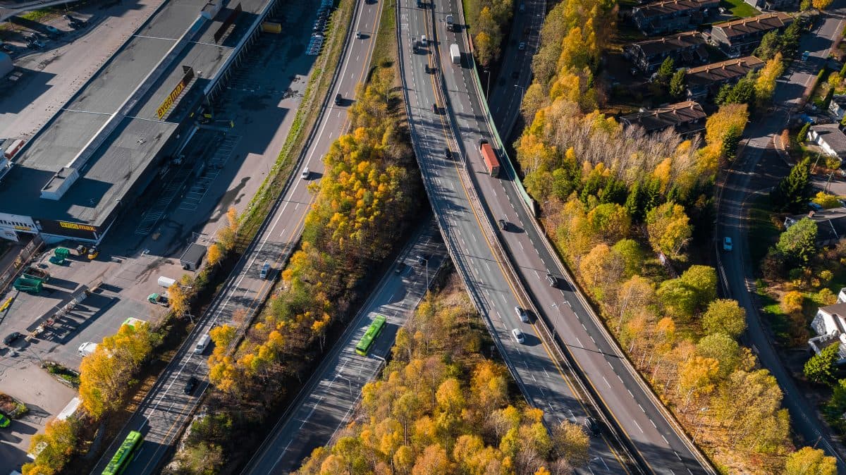 Flyfoto av kryssende motorveier med kjøretøyer, omgitt av høsttrær, ved siden av en jernbanelinje og et boligområde.